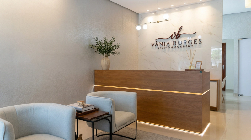 Recepção com balcão em Louro Freijó, parede em Mármore Branco e iluminação LED difusa, com poltronas claras.